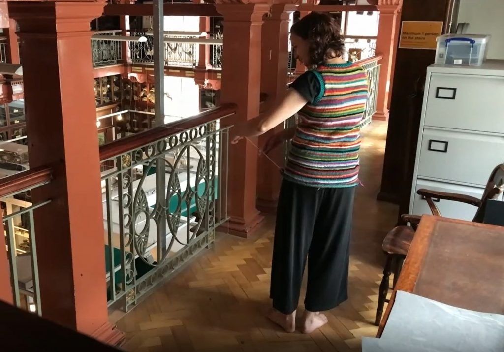 A woman with her back to the camera unravels yarn from the knitted jumper she is wearing and passes the end over a balcony. Louise Klarnett dances for _Unravelling_ (2021), performance stills.