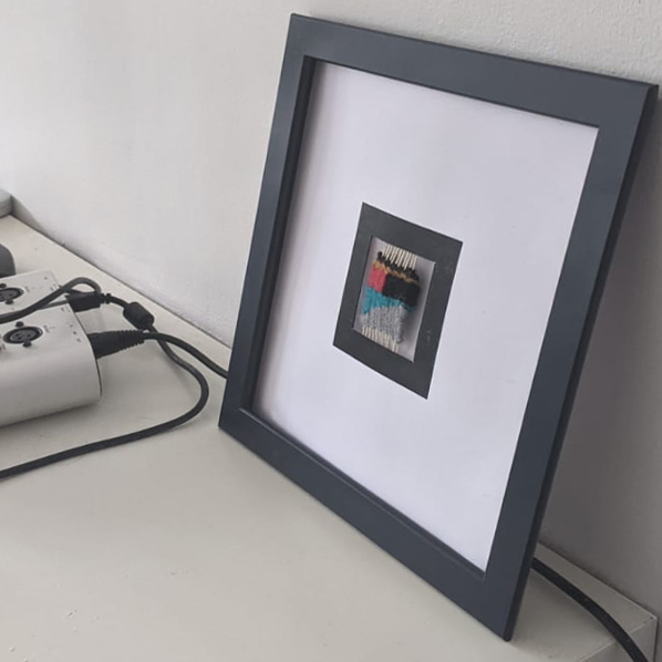 A square, blue picture frame leans against a wall. Inside the frame is a card cutout with another printed black square frame around a small woven marker.