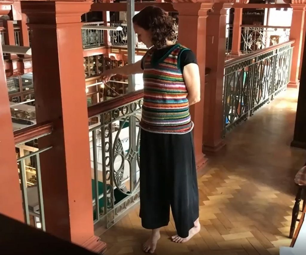 A woman facing towards the camera unravels yarn from the stripy knitted jumper she is wearing and passes the end over a balcony.