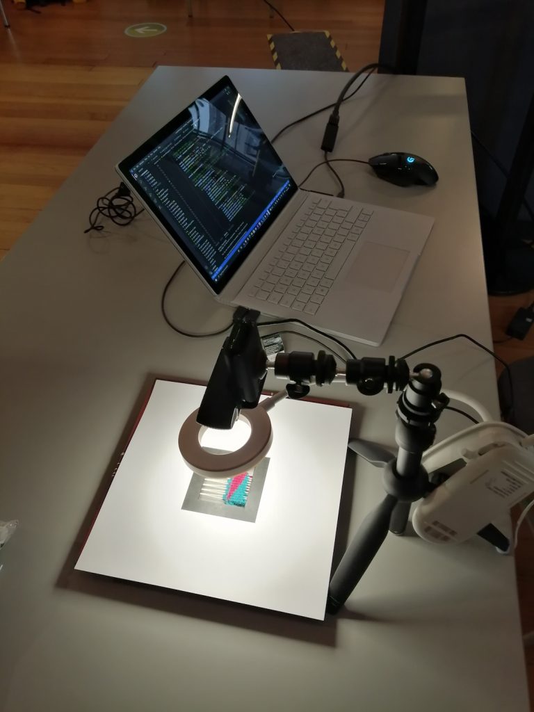 A web cam on an armature points downwards through a circle light to a woven sample beneath cutout card with a black square frame. The webcam is connected to an open laptop showing a page of code. 