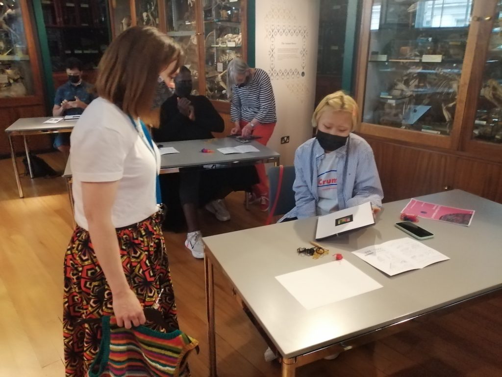 A seated woman shows her completed woven marker to another women standing to her right hand side. Both women wear face masks. In the background are more participants and the Grant Museum display cases.